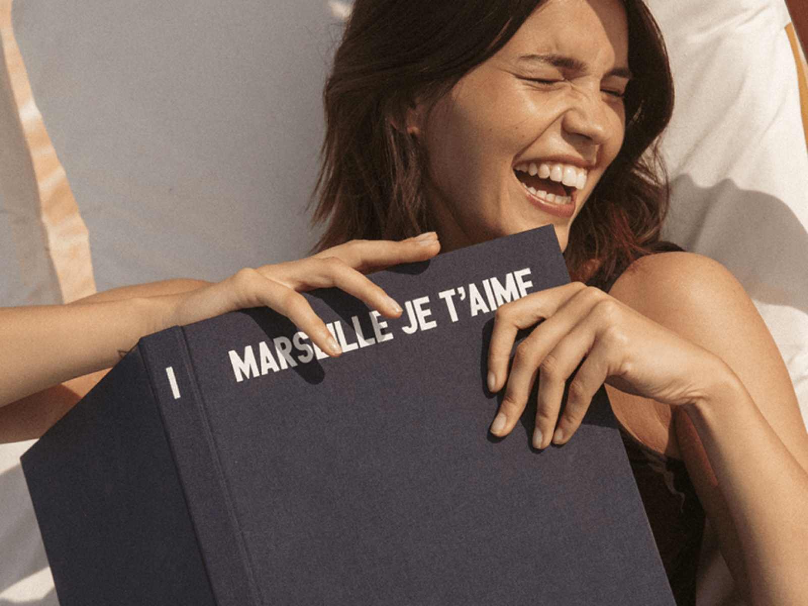 A woman smiles joyfully while holding a large book titled "Marsaille Je T'aime," laying on a sunbed