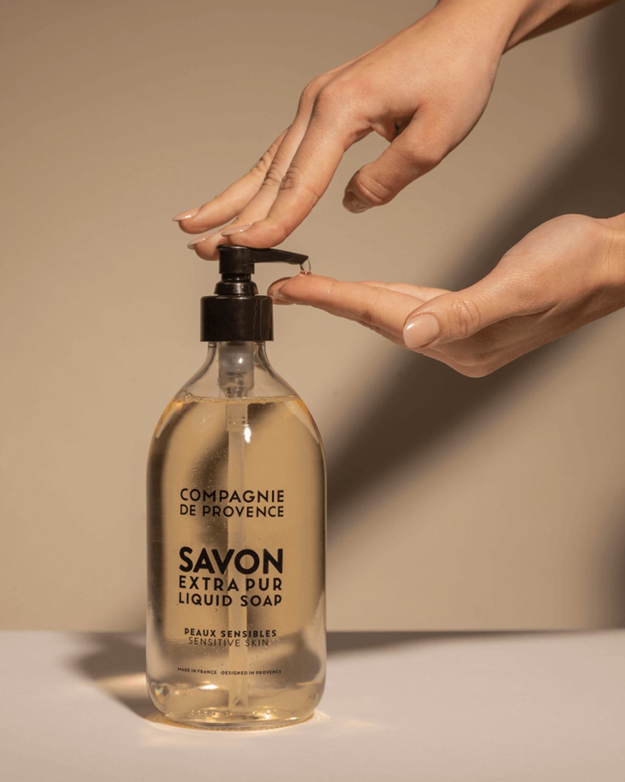 A hand is pressing down on the pump of a bottle of "Savon Extra Pur" liquid soap, which is clear and sits on a neutral background. The bottle features a black label with the brand name "Compagnie de Provence."