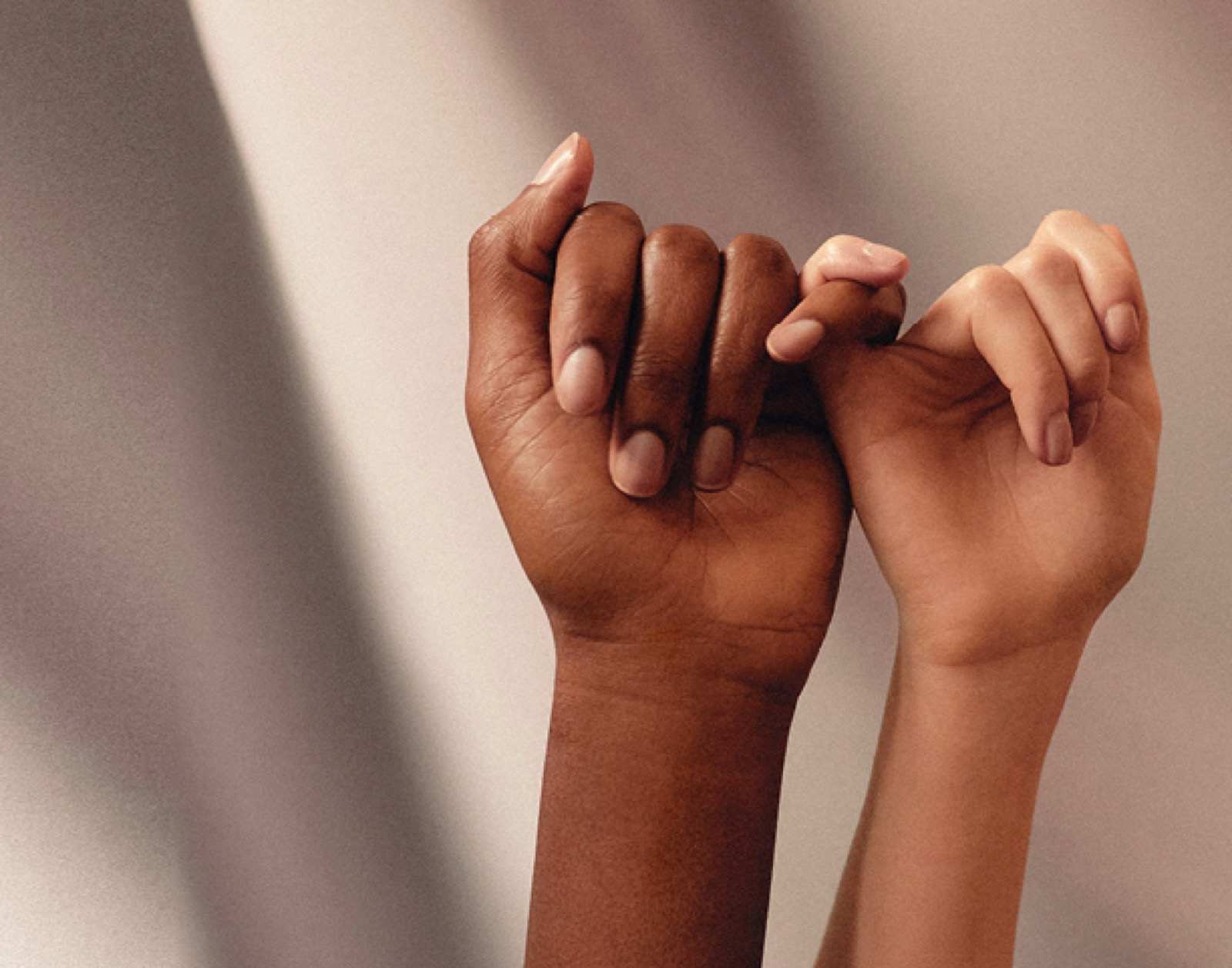 Two hands, one with darker skin and the other with lighter skin, are linked together in a pinky swear gesture against a softly lit background